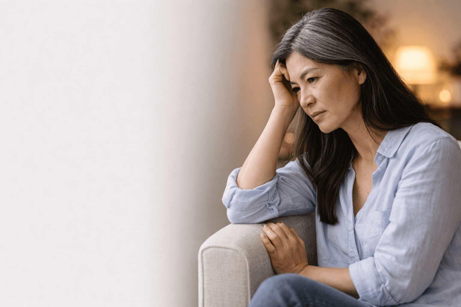 Pensive middle-aged Asian woman with grey-streaked hair sits on a sofa, leaning her head.