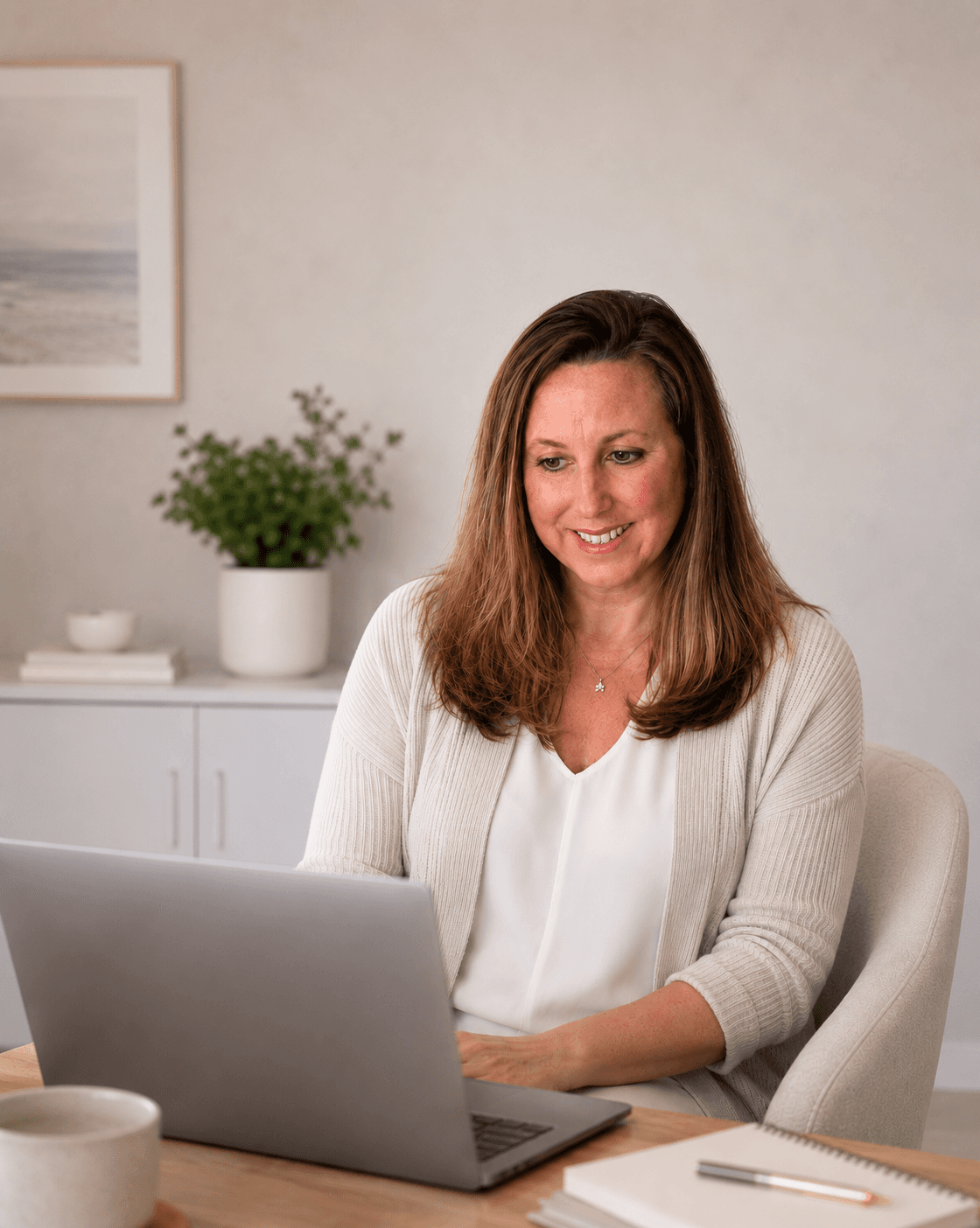 Smiling woman in a white cardigan works on a laptop at a bright, modern desk.