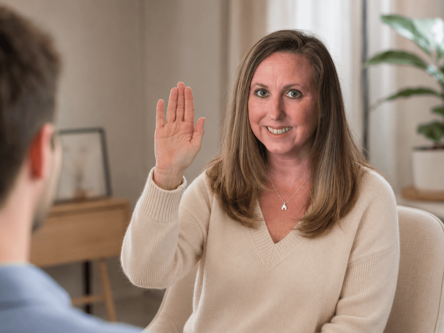 Smiling woman in a cream sweater raising her right hand while looking at another person.
