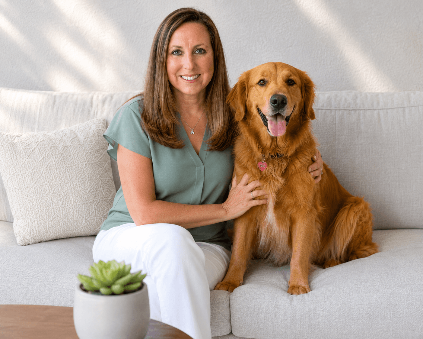 Smiling woman in green top sits on a couch with her happy golden retriever.