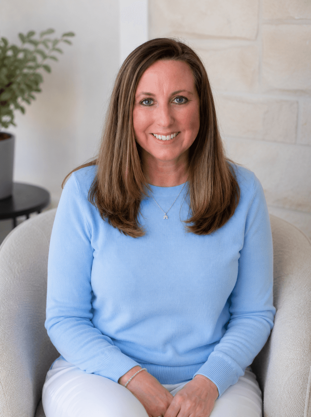 Smiling woman with long brown hair wearing a light blue sweater, seated in a beige armchair.