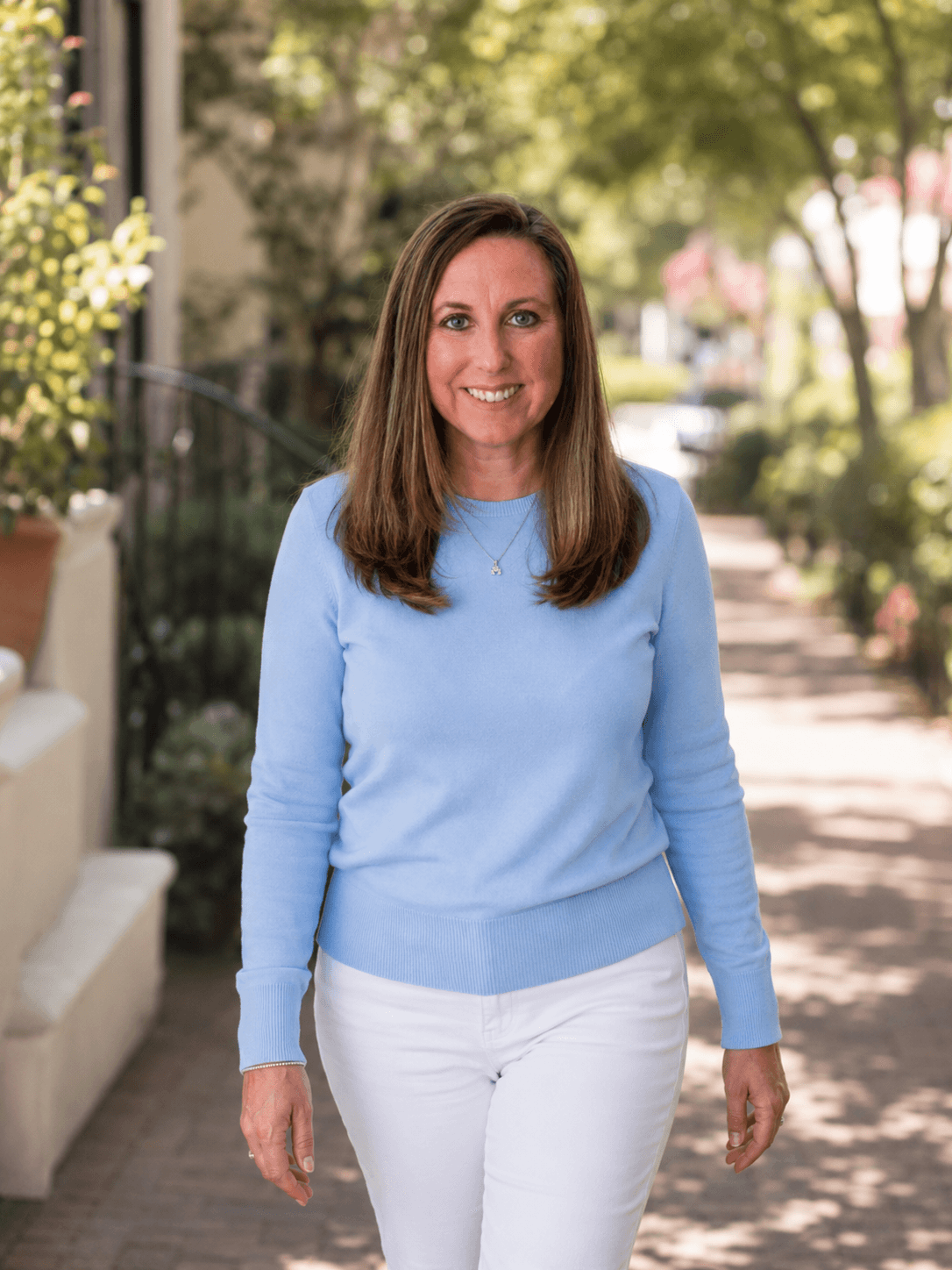 Smiling woman in a blue sweater and white pants walking on a sunlit brick path.