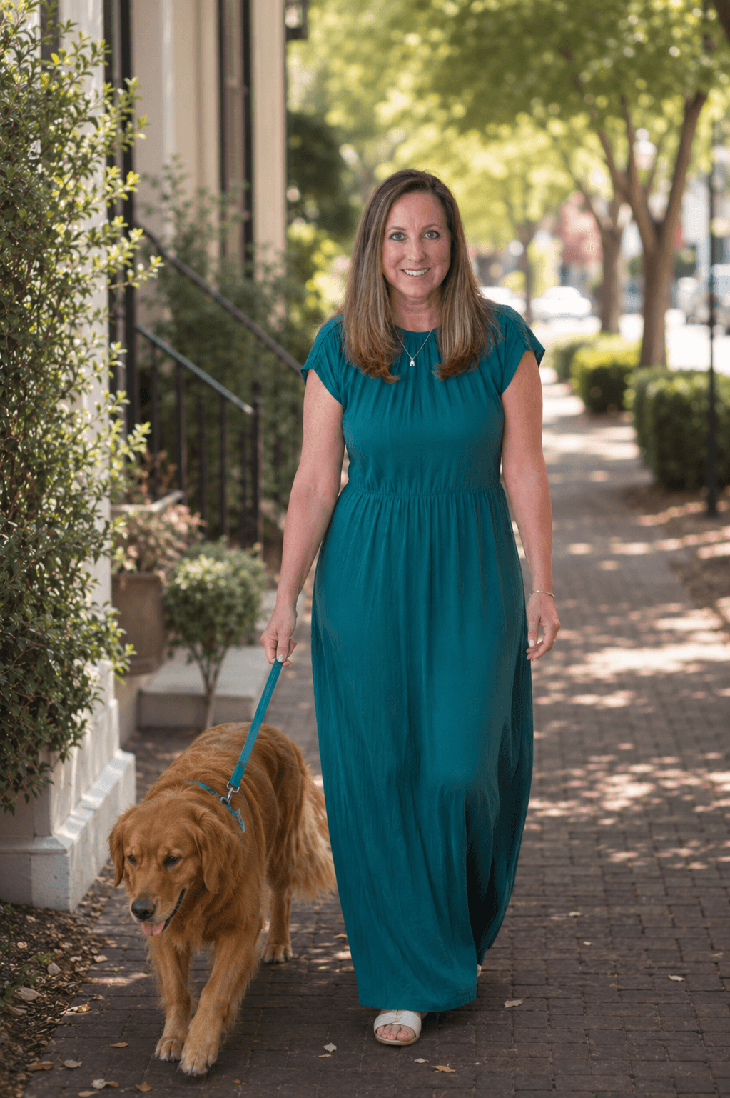 Smiling woman in a teal dress walks her golden retriever on a brick sidewalk.