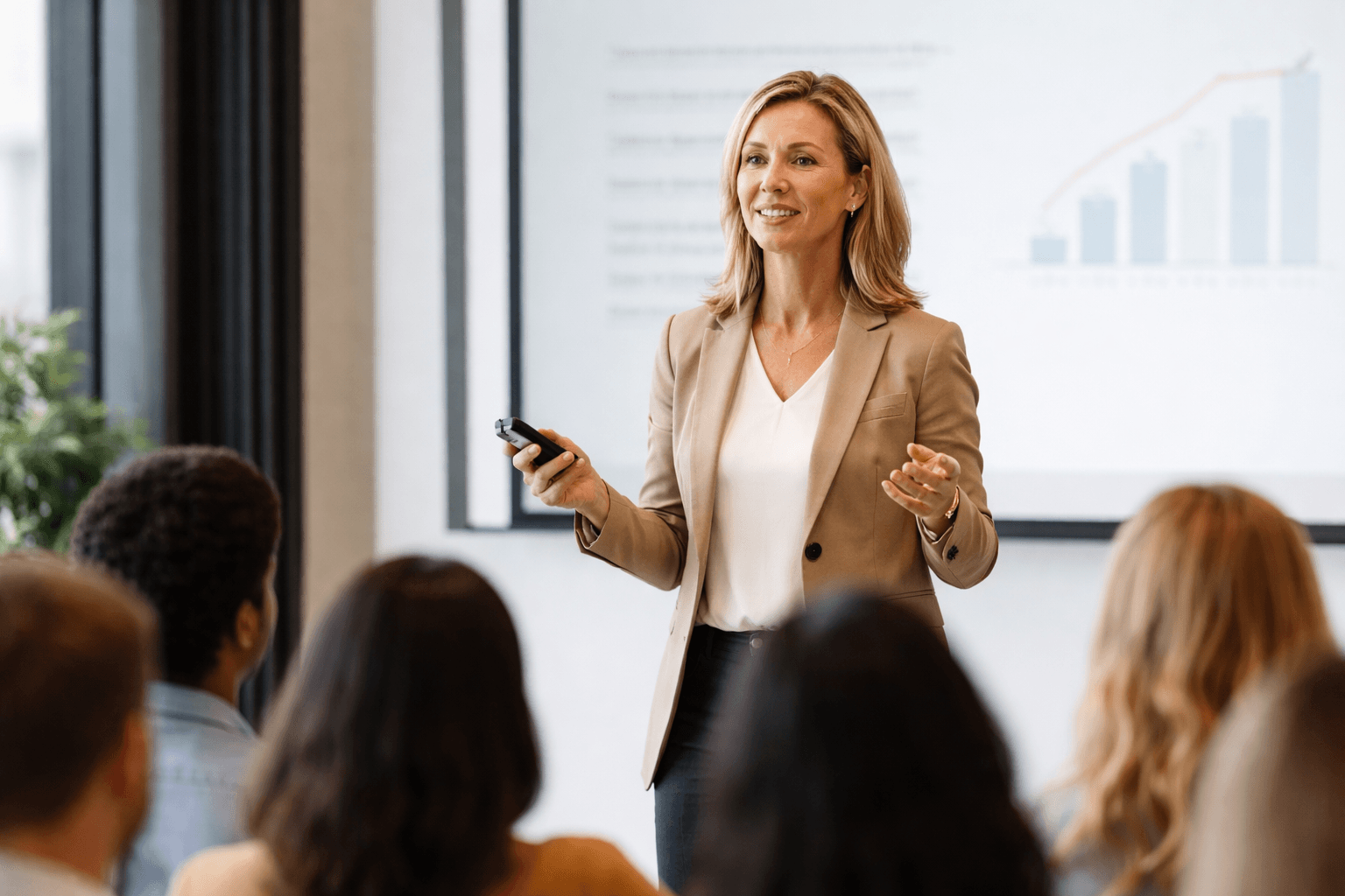 Professional woman in a tan blazer giving a presentation to an audience with charts.
