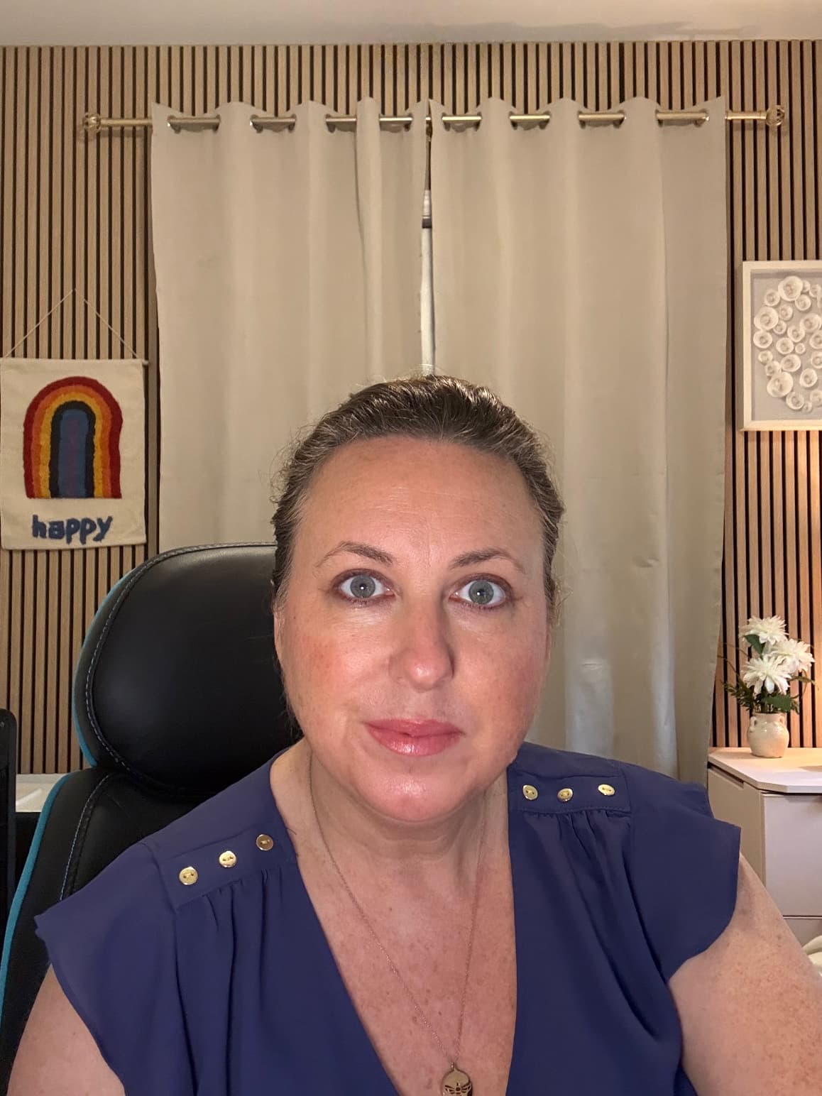 Woman with blue eyes in a blue top sitting before beige curtains and wood-slatted wall.