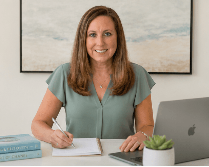 Smiling woman in green blouse writing in a notebook at a desk with laptop.