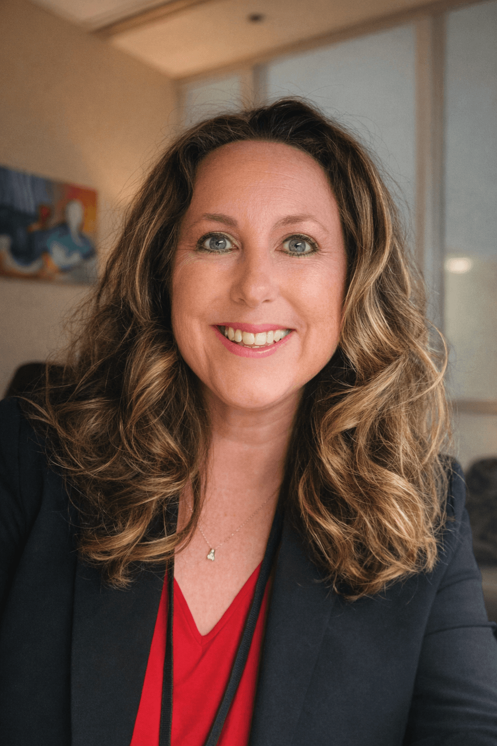 Smiling woman with wavy brown hair and blue eyes, wearing a black blazer and red top.